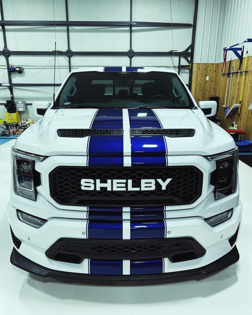 Front view of a white Shelby truck with blue racing stripes, sitting in a bay at the Flawless Finishes detailing shop.